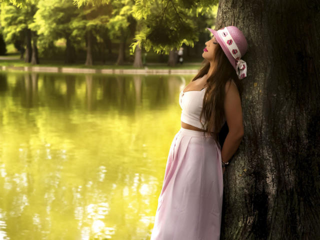 DashingFoxyX leans against a tree, wearing a white crop top and pink skirt, with a pink hat, showing her tits and ass, by a yellow-green reflecting pond.