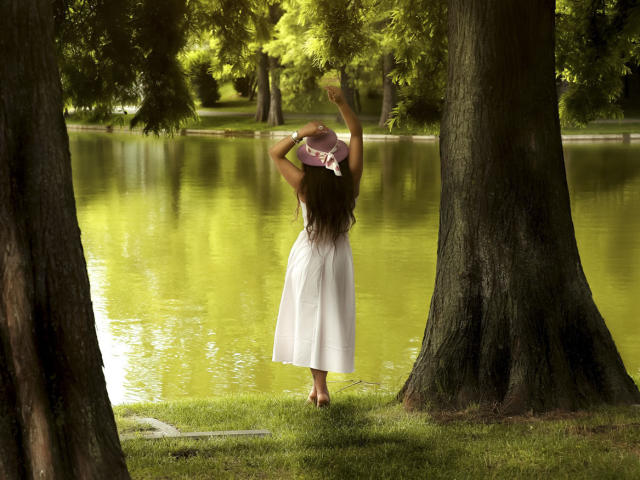 DashingFoxyX stands barefoot in a white dress, arms raised, removing a pink hat, back to camera, by a green-reflecting pond, flanked by tall trees.