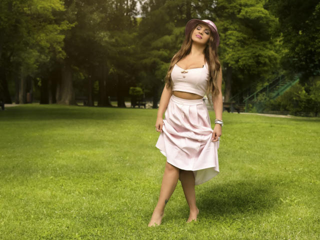 DashingFoxyX stands barefoot on green grass, wearing a white crop top, pink skirt, and purple hat, with long brown hair, small tits, and a sexy smile.