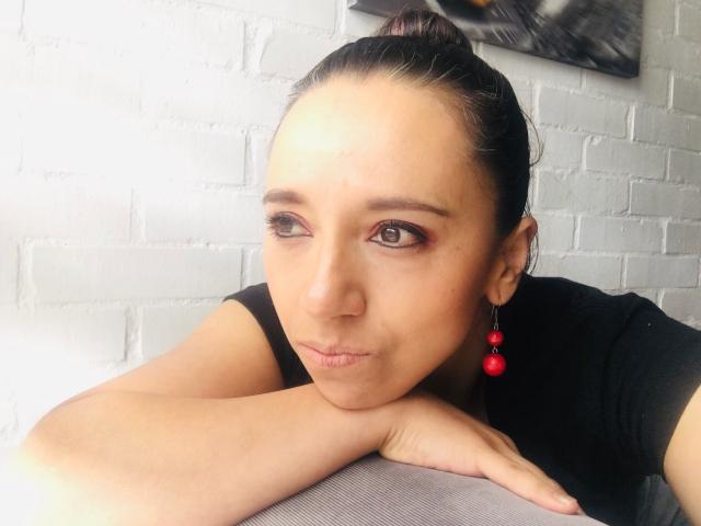 BellaLolita with tan skin, brown eyes, and red earrings, resting her chin on a gray surface, wearing a black shirt, smiling slightly. Her hair is in a bun.