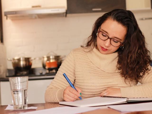 BellaLolita with curly brown hair, glasses, and a beige turtleneck, writing with a blue pen at a kitchen table, has a glass of water in front of her.