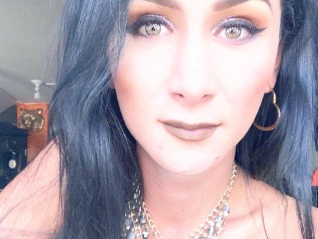 HugeCockTSforyou ha i capelli blu scuro, ombretto giallo-arancio, eyeliner nero, rossetto marrone, orecchini a cerchio dorati e una collana a catena d'argento, con un'espressione leggermente seria..