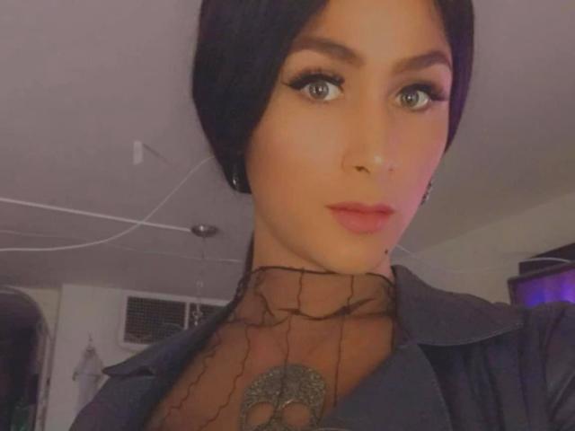 HugeCockTSforyou, con capelli neri, occhi azzurri e rossetto rosa, indossa una collana nera e una giacca di pelle, e ha un tatuaggio sul collo. L'illuminazione è soffusa e calda..
