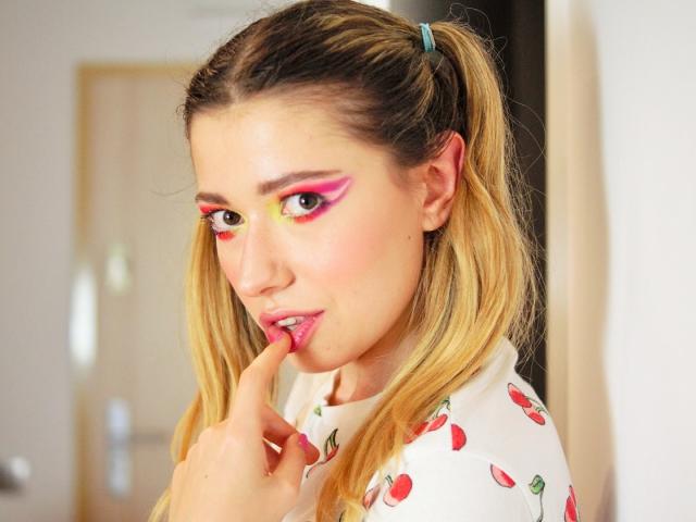 HeartHeidi, avec ses couettes blondes, son maquillage ocre et jaune, et sa chemise à motifs cerises, mordant son ongle avec du rouge à lèvres rose, regardant l'appareil photo avec une expression séduisante..