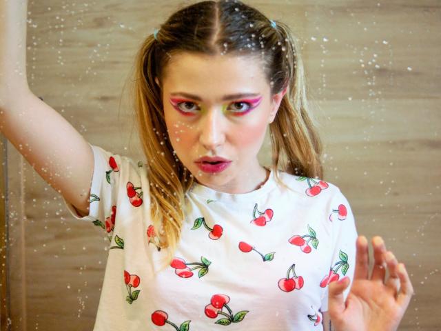 HeartHeidi with puffy pink eyeliner, white cherry print shirt, blonde pigtails, and lipstick, standing in front of a dirty glass window, looking up with slightly parted lips.