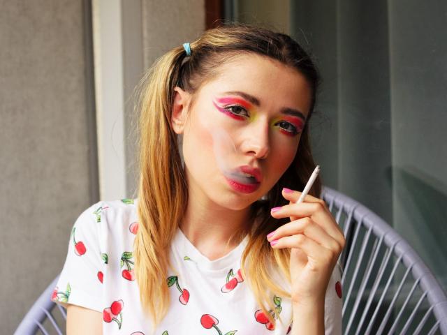 HeartHeidi with blonde pigtails, wearing a cherry print shirt, has bright pink and yellow eyeshadow, gray lipstick, and holds a cigarette. She's sitting in a gray chair.