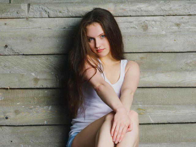 Ariadna, met lang bruin haar, draagt een wit tanktopje en blauwe korte broek, zittend tegen een houten muur, benen gekruist, armen om de knieën, met een lichte glimlach, kijkend naar de camera..