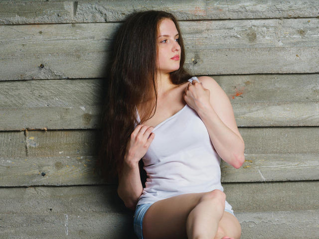 Ariadna, con lunghi capelli castani, una canotta bianca e pantaloncini blu, seduta contro un muro di legno, tiene la canotta con una mano e guarda di lato..