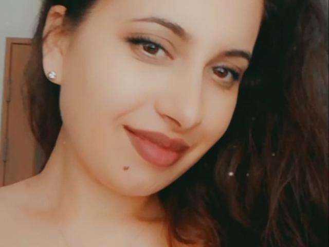 Il viso di SweetJeniffer, capelli castani ondulati, sopracciglia scure, sorriso delicato, occhi castani, rossetto scuro, una piccola lentiggine sotto il labbro, indossa orecchini di diamanti, pelle liscia, leggero rossore..