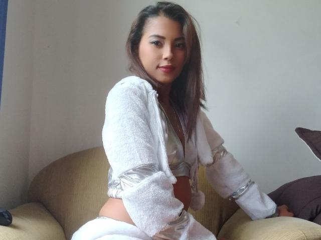 KolorBella has light brown skin, brown hair, and wears a white, fluffy, cropped, long-sleeve top with a shiny silver bra, sitting on a beige couch, smiling.