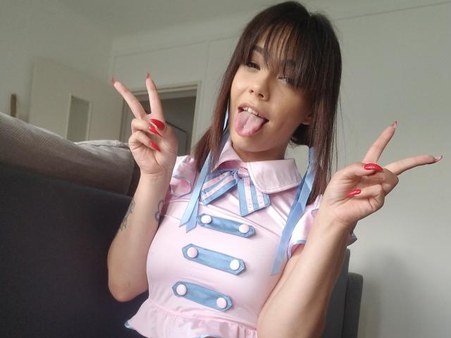 KassiKassou, com cabelo castanho, vestido rosa, fitas azuis e botões, de língua para fora, fazendo dois sinais de paz, unhas vermelhas, pequena tatuagem no braço esquerdo..