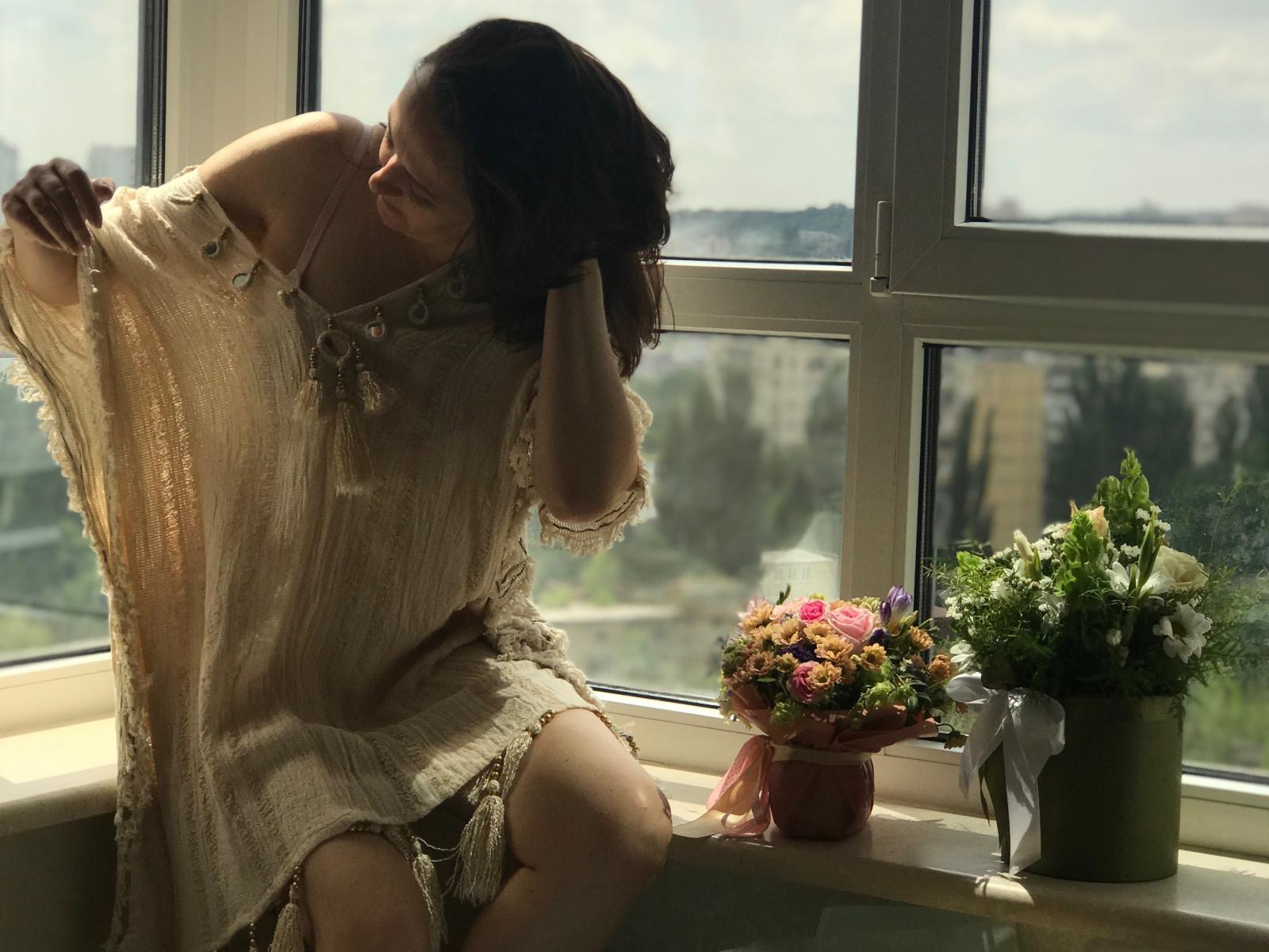 UllimOhi sits by a window, wearing a sheer, beige dress, pulling it up to expose her ass. She has glasses and dark hair, looking at flowers and a silver cat.