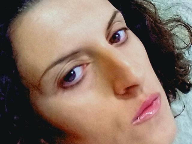 Audrey Daisys Gesicht, mit ihren braunen, lockigen Haaren, violetten Augen und rosafarbenen Lippen, zeigt einen leicht gelangweilten oder müden Ausdruck. Ihre Haut ist hell und glatt..