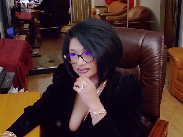 ClassybutNaughty, con cabello negro, gafas y ojos morados, vestida con un atuendo negro, tiene un dedo en los labios, mostrando un anillo y una pulsera, sentada en una silla de cuero..