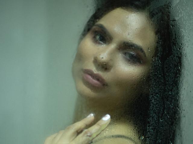 BrianaPreston's face is pressed against a foggy window with droplets, her dark curly hair and sultry eyes looking slightly to the side, her hand near her chin.