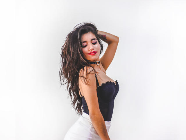 CrystalJoness, com longos cabelos negros ondulados, vestindo um corpete de renda preta, saia branca e batom vermelho, posando com o braço levantado, exibindo seus seios e uma tatuagem no braço..