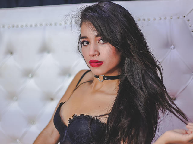 CrystalJoness with long black hair, red lipstick, black lace bra, and a black choker, looking to the side, sitting on a white, tufted, diamond-studded sofa.