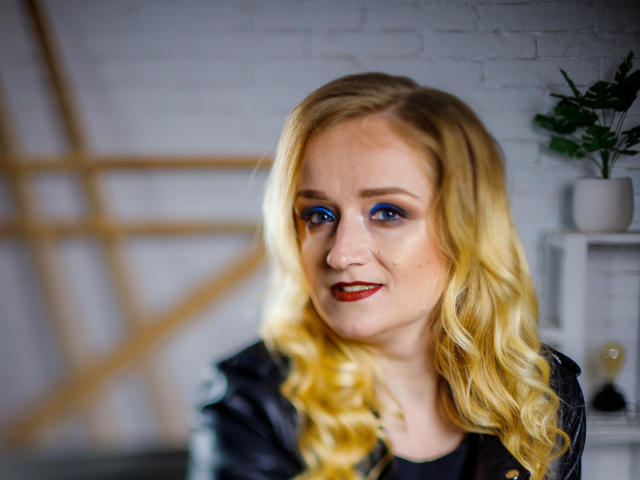 OhMollyHoney with blonde curls, blue eye makeup, red lipstick, wearing a black shirt, looking slightly to the side with a subtle smile, in a blurred indoor setting.