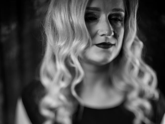Black-and-white photo of OhMollyHoney with wavy blonde hair, dark lipstick, and a subtle smile. Her eyes are half-closed, and she's wearing a dark top.