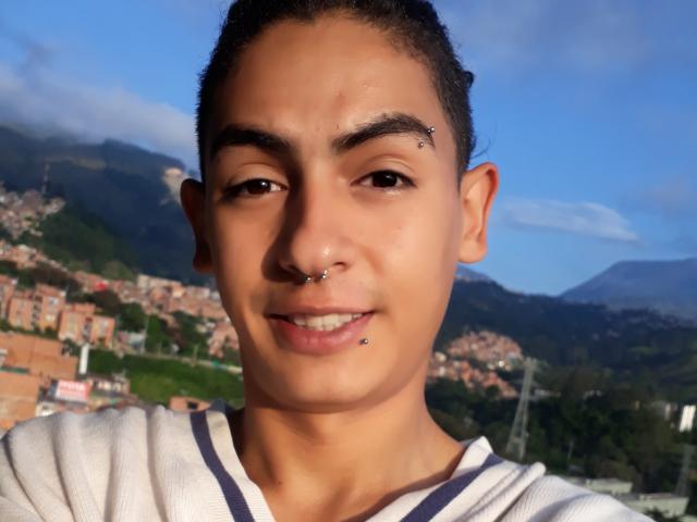 AlexanderBoy ha un viso carino con occhi marrone scuro, capelli neri corti e indossa una camicia bianca con strisce scure. Ha un piercing al naso e un piercing al labbro inferiore..