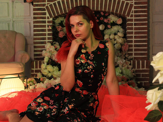 MelanyTessk with red hair, floral dress, and red tulle, sitting with hand near mouth, looking at camera, showing her tits, in a cozy, floral-filled setting.