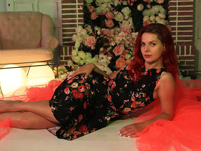 MelanyTessk with red hair, wearing a black floral dress, lying on a red tulle, looking at the camera, with her ass and tits partially visible, in a dimly lit room.