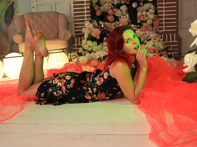 MelanyTessk lies on a white floor, wearing a black floral dress, with red hair and green highlights, legs up, hands near mouth, surrounded by red fabric and white flowers.