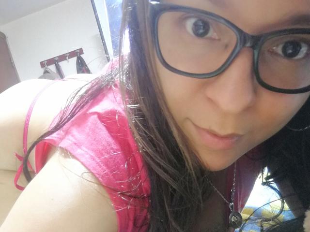 Angelonix with glasses, pink shirt, and red thong, bent over, showing her ass, looking at the camera with a slight smile, dark wet hair, and a necklace.