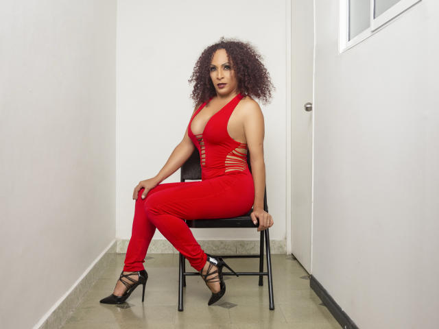 KhazandraX with curly brown hair, red cutout dress, showing tits, sitting on black chair, wearing black strappy heels, in white-walled room. Her ass is slightly visible.