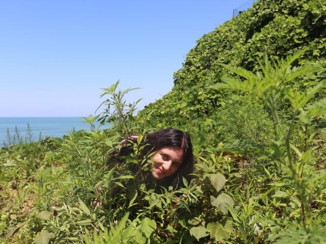 RoksanaBest está agachada em meio à folhagem verde, com o rosto parcialmente escondido, tendo cabelos pretos e pele clara, sob um céu azul brilhante, com um mar calmo ao fundo..