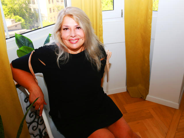ByaStorm, blonde, souriante, robe noire, assise, rideaux jaunes, fenêtre, plante verte, sol en bois, murs blancs, bras sur la chaise, main sur la plante, aux formes généreuses, mignonne, poitrine moyenne..