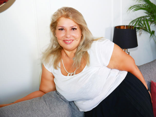 ByaStorm, blonde, medium tits, white shirt, black skirt, necklace, smiling, leaning on gray couch, black lamp in background, green plant to the right.