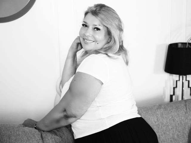 ByaStorm, une femme aux formes généreuses avec des cheveux blonds, souriant à l'appareil photo, portant un haut blanc et une jupe noire, assise sur un canapé gris, regardant par-dessus son épaule..