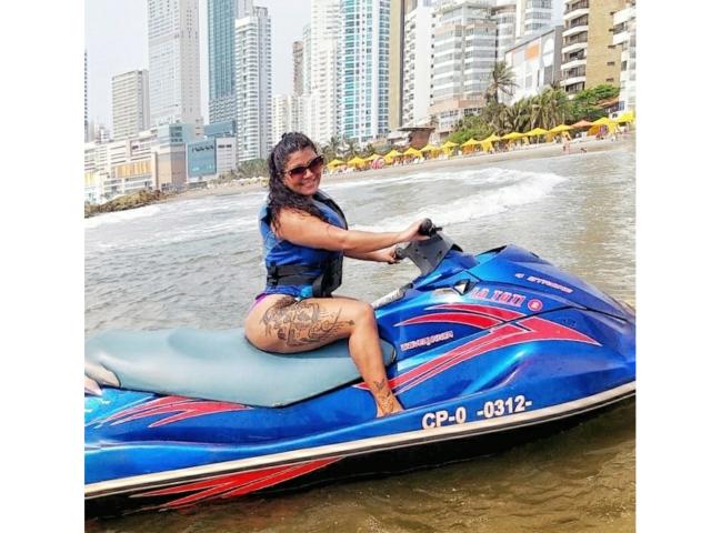 CanelleBom mit Tattoos, trägt eine blaue Schwimmweste, fährt auf einem blau-roten Jetski, sitzt auf dem Wasser, lächelt, trägt eine Sonnenbrille, im Hintergrund Strand und hohe Gebäude..