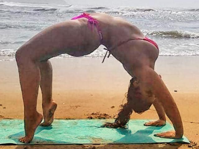 CanelleBom está inclinada sobre una esterilla verde, vestida con un bikini rosa, con las nalgas levantadas y la cabeza hacia abajo, en una playa arenosa con suaves olas de fondo..