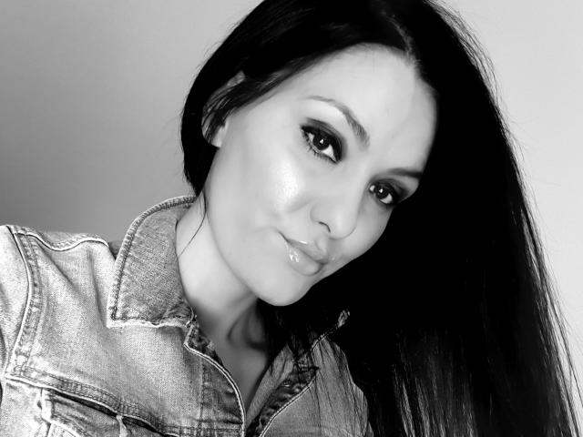 Selfie em preto e branco de JoliDanielle com lábios brilhantes, vestindo uma jaqueta jeans, inclinada para frente, com cabelo preto longo e um leve sorriso, mostrando seus seios de forma discreta na imagem..