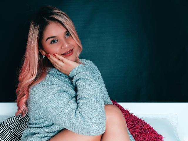 ValerieStone with pink hair, gray knit sweater, red nails, smiling, hands on cheeks, sitting on white and red pillows, dark background. She's cute with a sexy, relaxed vibe.