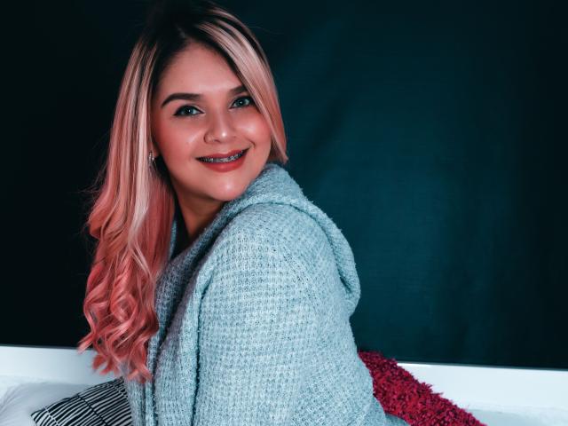 ValerieStone with pink and blonde hair, wearing a gray knitted hoodie, smiling at the camera, showing her cute face and small nose, sitting on a white and red surface.