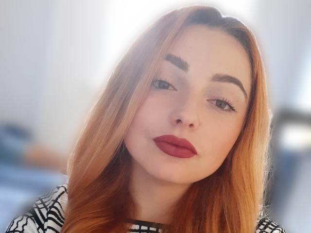 KellyMareta, avec ses longs cheveux orange, ses lèvres rouges pulpeuses et ses sourcils épais, regarde l'appareil photo avec un léger moue. Elle porte un haut rayé noir et blanc..