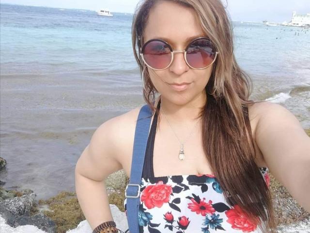 Angelonix with long brown hair, wearing pink-tinted sunglasses, floral dress, and blue suspenders, has a small, cute nose, and is pouting slightly at the beach.