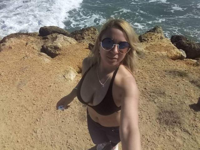 Angelonix with blonde hair, wearing sunglasses and a black bikini top, showing her tits, standing on a rocky beach with ocean waves in the background, taking a selfie.