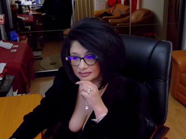 "ClassybutNaughty", con cabello oscuro, gafas y sombra de ojos morada, vestida con una blusa negra, está sentada en una silla de cuero, sosteniendo un anillo y sonriendo con lápiz labial rojo..