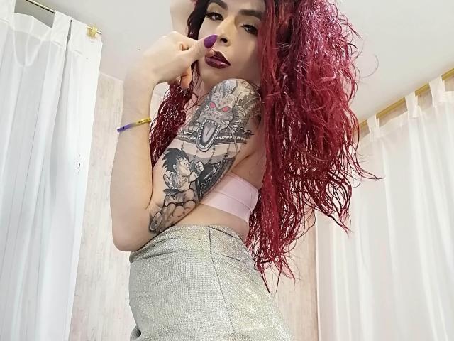 Rosariio, avec ses cheveux roux bouclés, son rouge à lèvres violet, son bras tatoué, portant un soutien-gorge rose et un pantalon argenté scintillant, tenant un rouge à lèvres violet, regardant la caméra, avec des rideaux blancs en arrière-plan..