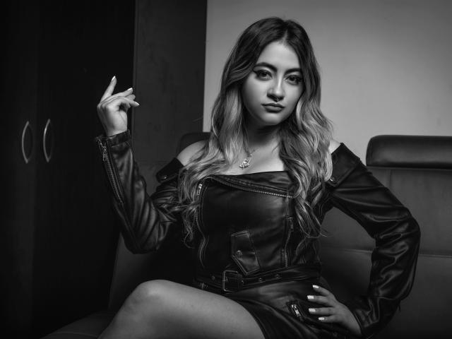 Black and white photo of ArleenHunter with long wavy hair, wearing a leather dress, sitting with one hand on her thigh and the other holding a cigarette. Her ass is visible.