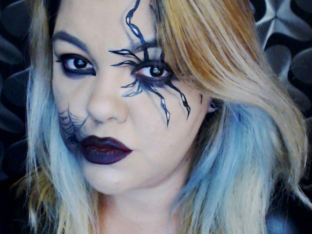 LaDominatrice has dark, spider-web tattoos around her eyes, purple lipstick, and blonde-blue hair. Her eyes are black with dramatic eyeliner, and she's looking directly at the camera.