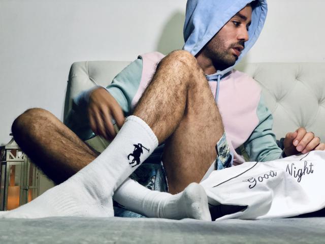 SionActif with hairy legs, wearing a blue hoodie, pink and green jacket, white socks with a black horse logo, sitting on a bed, looking to the side, holding a shirt.
