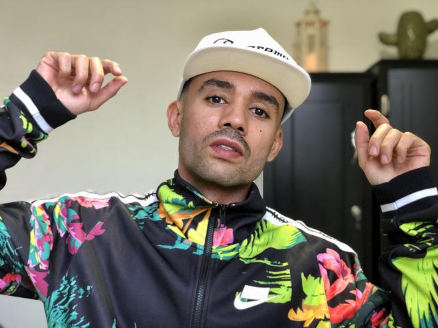 SionActif with tan skin, dark eyebrows, and a goatee, wearing a white cap and colorful Nike jacket, holding his hands up. His jacket has bright, abstract patterns.