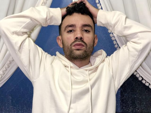 SionActif with hands in dark hair, wearing white hoodie, blue background, slightly dirty, short beard, serious expression, medium skin, dark eyebrows, small nose, small mustache.