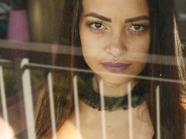 AmaraLove's face through a window, nose ring, purple lipstick, dark brown eyes, straight brown hair, black choker, looking directly at camera, sunlight, metal bars in foreground.