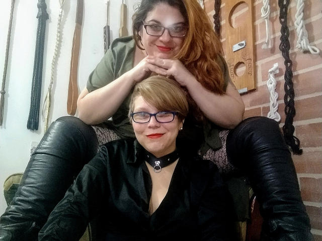 Two LatinoHardGirls with glasses, one with red hair and the other with blonde, sitting in a room filled with BDSM gear. One has black boots and the other's tits are exposed.